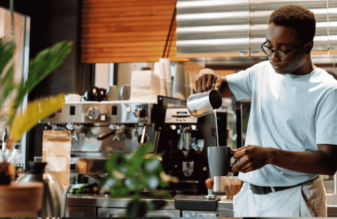 A barista wearing glasses pours steamed milk into a cup at a coffee shop, standing in front of an espresso machine and surrounded by plants and coffee equipment, while wrapped in the cozy comfort of a winter blanket.