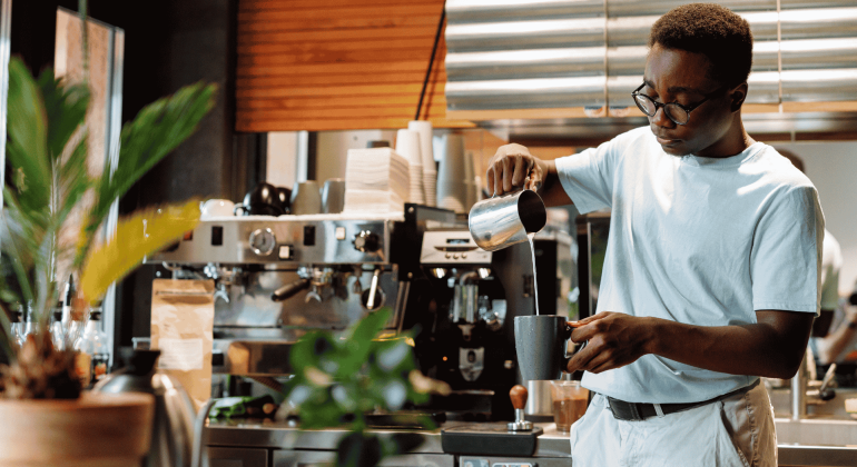 A barista wearing glasses pours steamed milk into a cup at a coffee shop, standing in front of an espresso machine and surrounded by plants and coffee equipment, while wrapped in the cozy comfort of a winter blanket.