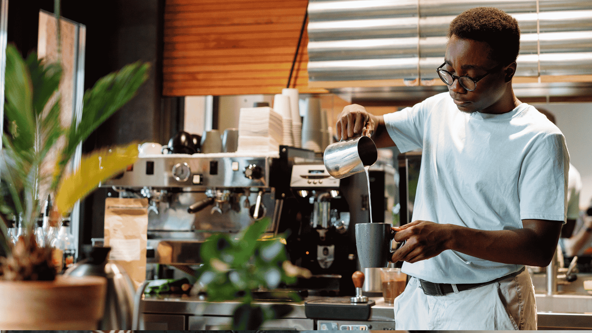 A barista wearing glasses pours steamed milk into a cup at a coffee shop, standing in front of an espresso machine and surrounded by plants and coffee equipment, while wrapped in the cozy comfort of a winter blanket.