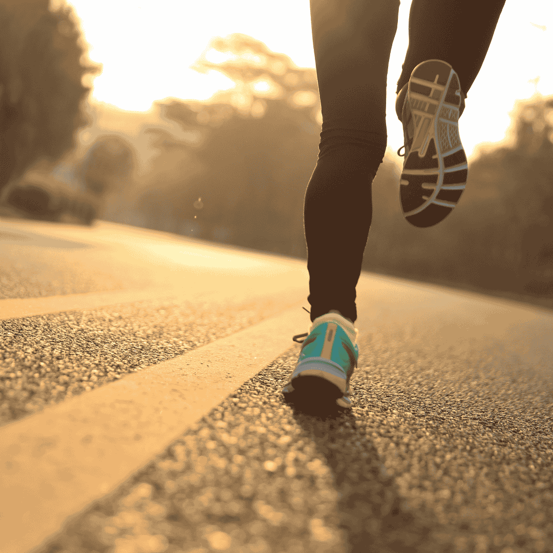 Close-up of a person jogging on an empty road at sunrise, wearing black leggings and blue running shoes. The sunlight creates a warm, golden glow and long shadows on the pavement, reminiscent of the comforting embrace of a winter blanket against the early morning chill.