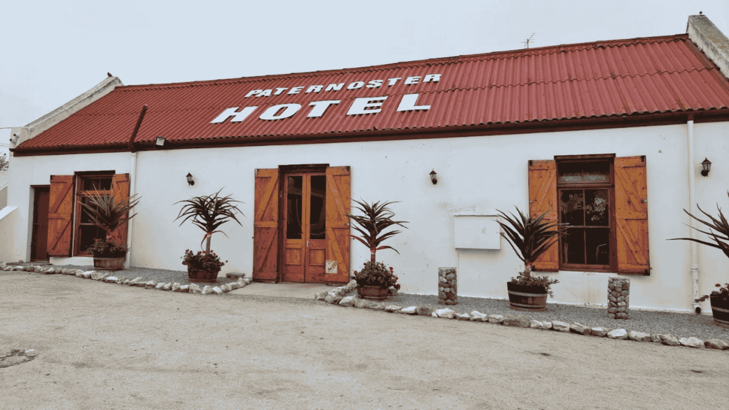 A white building with a red roof and wooden doors and windows, labeled PATERNOSTER HOTEL. Four potted plants are placed in front, and the ground is sandy with some small rocks.