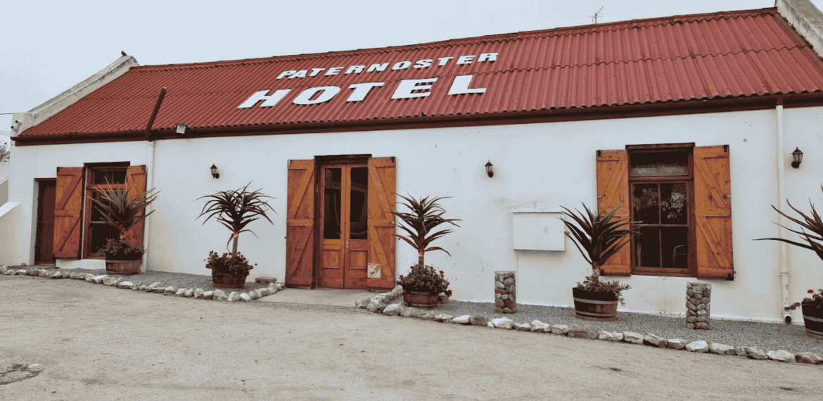 A white building with a red roof and wooden doors and windows, labeled PATERNOSTER HOTEL. Four potted plants are placed in front, and the ground is sandy with some small rocks.