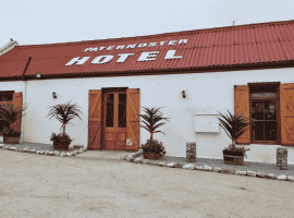 A white building with a red roof and wooden doors and windows, labeled PATERNOSTER HOTEL. Four potted plants are placed in front, and the ground is sandy with some small rocks.