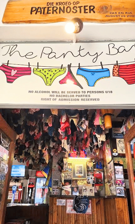 A bar entrance with a sign reading The Panty Bar, decorated with drawings and real hanging underwear, and a notice restricting alcohol to under-18s and bachelor parties. Walls display posters and memorabilia.