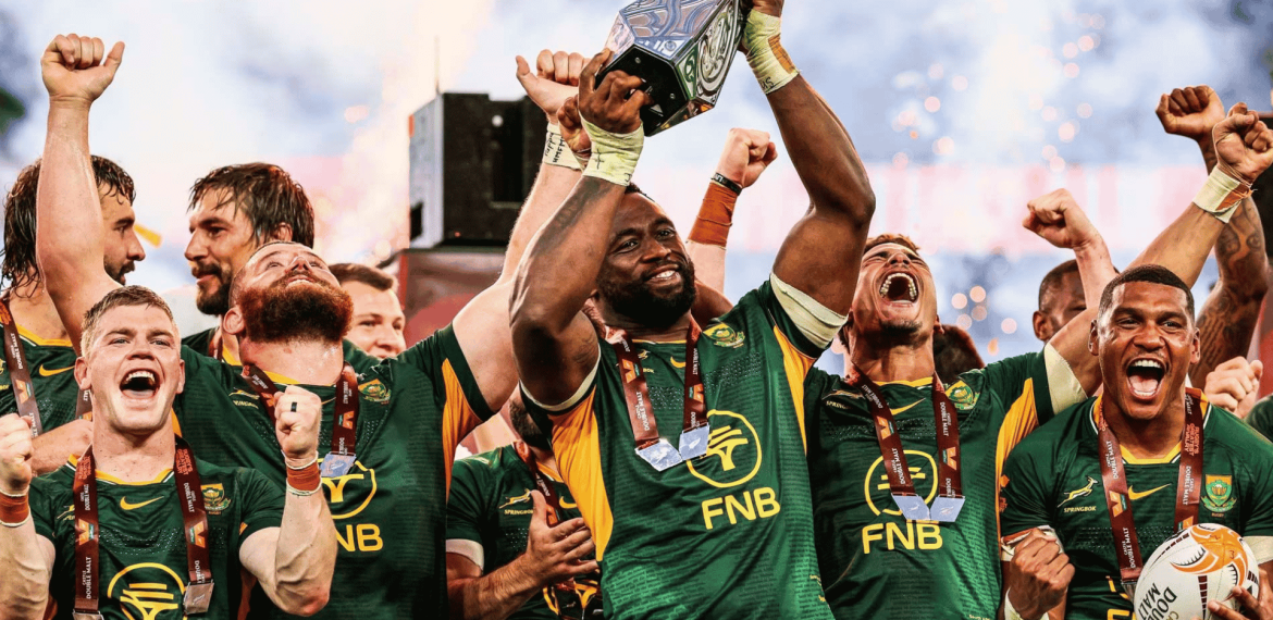 South African rugby players celebrate joyfully, wrapped in green jerseys and medals like a winter blanket, as their captain lifts a trophy with fireworks in the background.