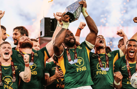 South African rugby players celebrate joyfully, wrapped in green jerseys and medals like a winter blanket, as their captain lifts a trophy with fireworks in the background.