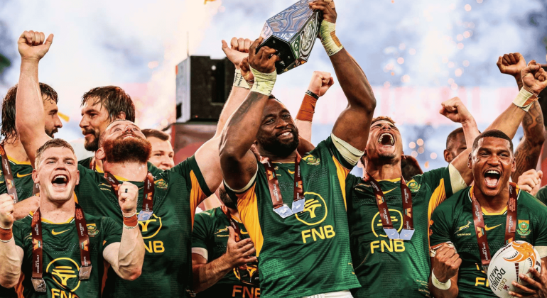 South African rugby players celebrate joyfully, wrapped in green jerseys and medals like a winter blanket, as their captain lifts a trophy with fireworks in the background.