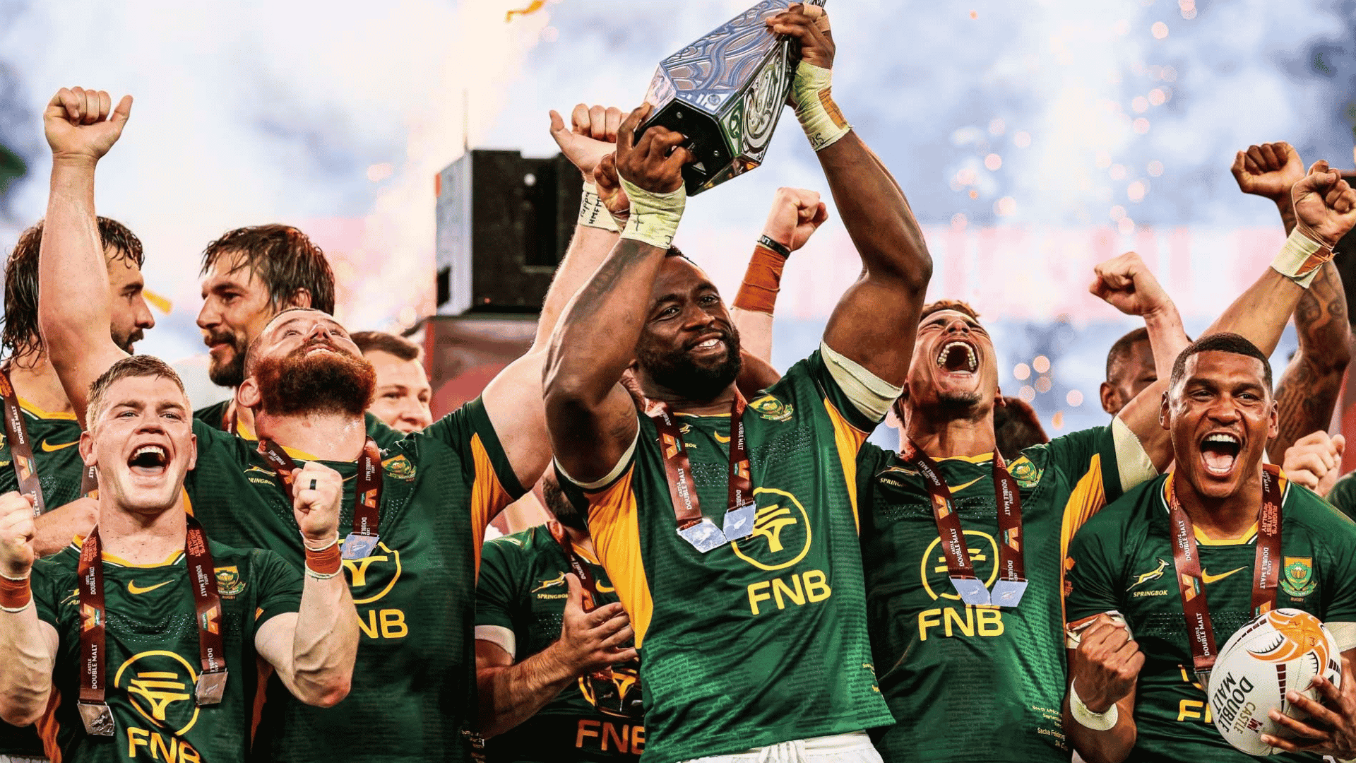 South African rugby players celebrate joyfully, wrapped in green jerseys and medals like a winter blanket, as their captain lifts a trophy with fireworks in the background.