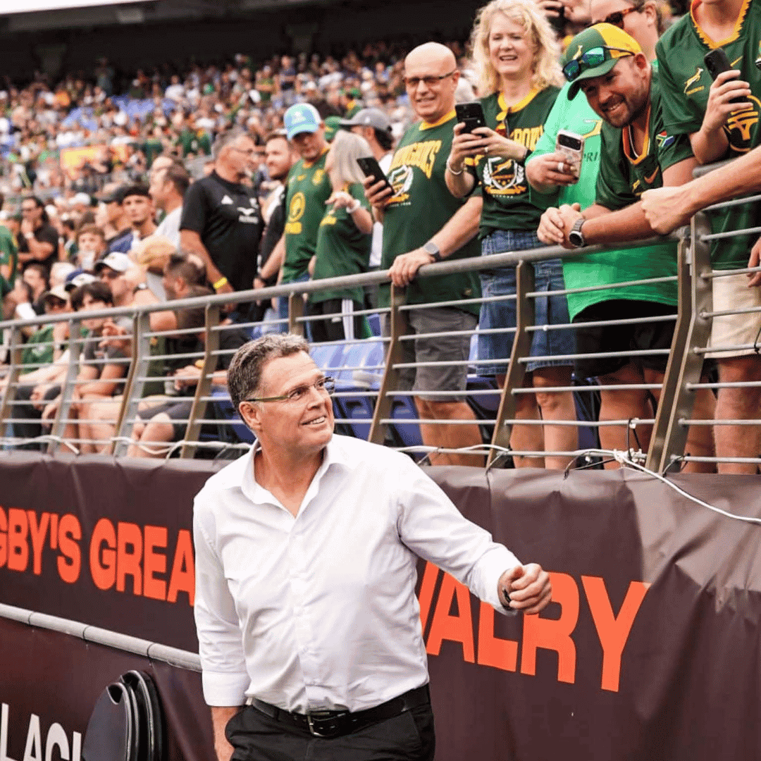 A man in a white shirt walks along a stadium barrier, smiling up at cheering fans in green jerseys. The stands are full, with a few spectators wrapped in winter blankets as they lean over the railing to interact with him.