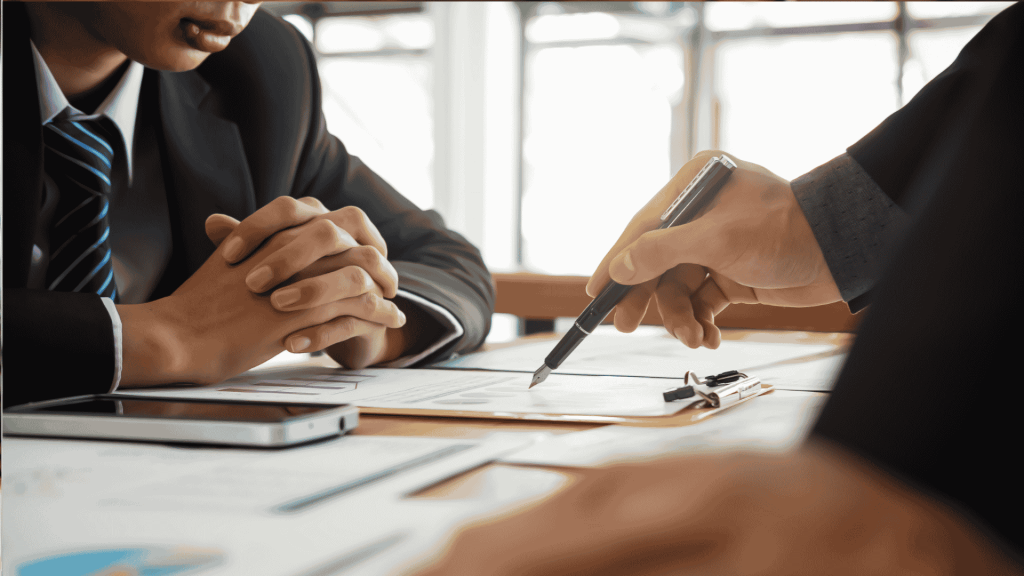 Two people in business suits sit at a desk with documents, a clipboard, and a smartphone, discussing paperwork as one person points with a pen. A cozy winter blanket draped over the back of one chair adds a touch of warmth to the professional setting.