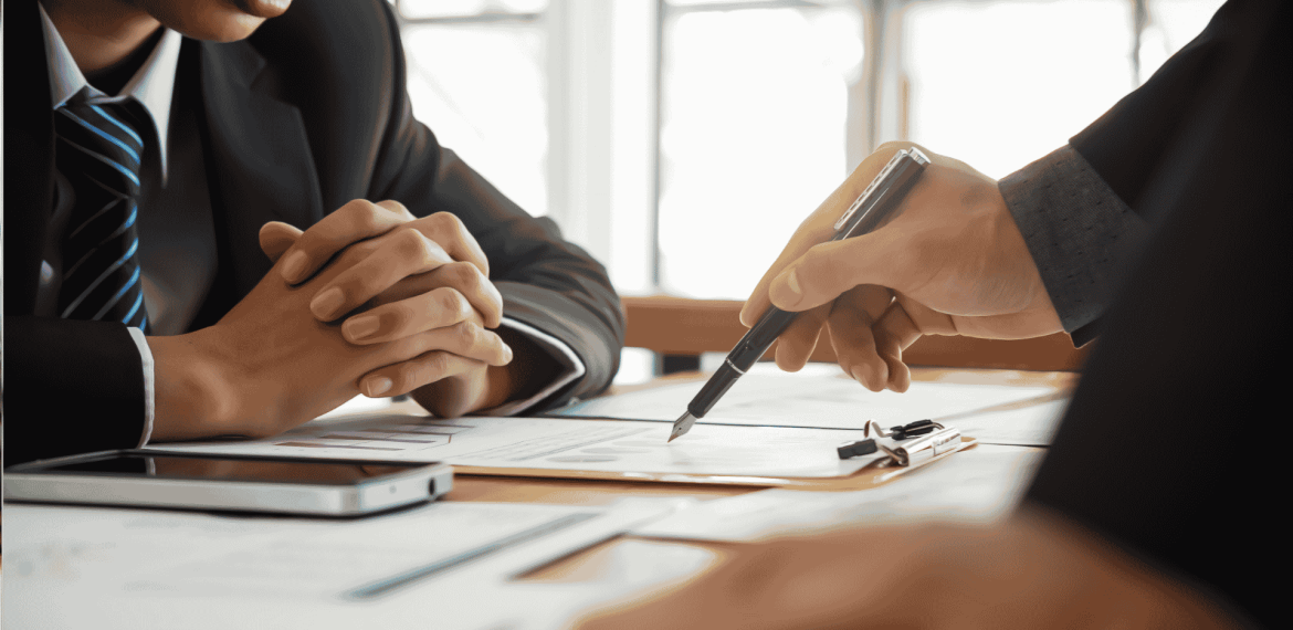 Two people in business suits sit at a desk with documents, a clipboard, and a smartphone, discussing paperwork as one person points with a pen. A cozy winter blanket draped over the back of one chair adds a touch of warmth to the professional setting.