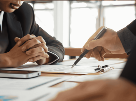 Two people in business suits sit at a desk with documents, a clipboard, and a smartphone, discussing paperwork as one person points with a pen. A cozy winter blanket draped over the back of one chair adds a touch of warmth to the professional setting.