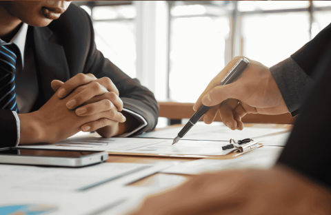 Two people in business suits sit at a desk with documents, a clipboard, and a smartphone, discussing paperwork as one person points with a pen. A cozy winter blanket draped over the back of one chair adds a touch of warmth to the professional setting.