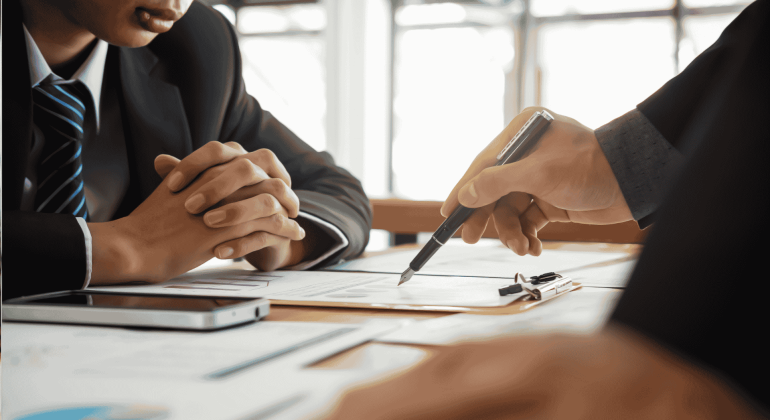 Two people in business suits sit at a desk with documents, a clipboard, and a smartphone, discussing paperwork as one person points with a pen. A cozy winter blanket draped over the back of one chair adds a touch of warmth to the professional setting.