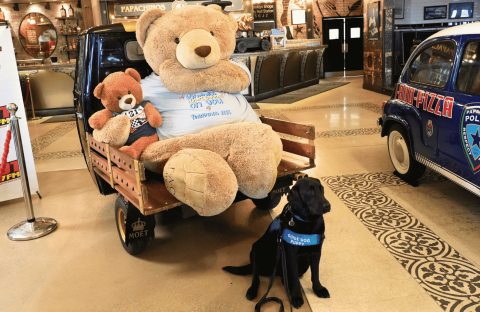 A large teddy bear and a small teddy bear sit on a cart, while a black service dog with a blue vest sits in front. Vintage cars and restaurant decor are visible in the background.