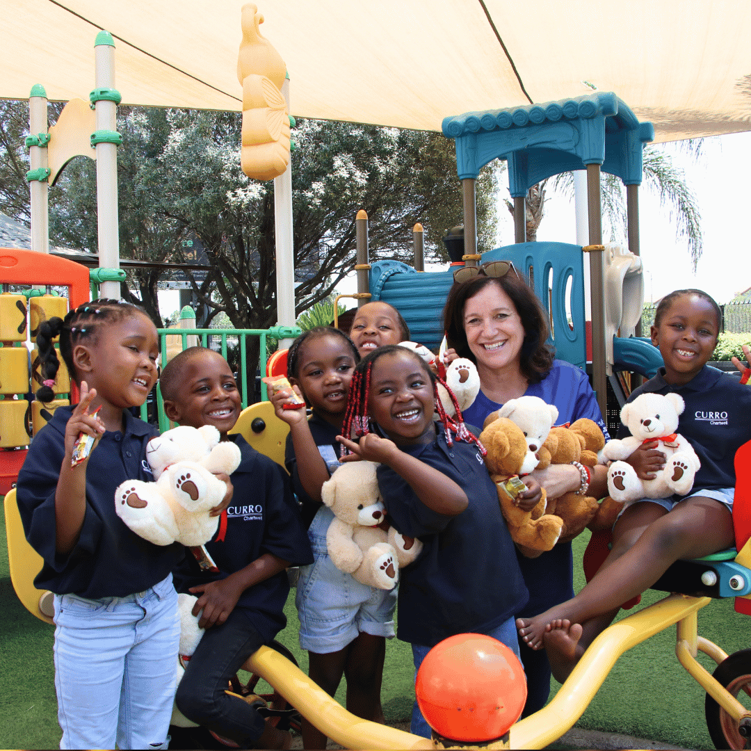 A group of smiling children and an adult sit on playground equipment, holding teddy bears. They are outside, surrounded by colorful play structures and greenery, enjoying a cheerful moment together.