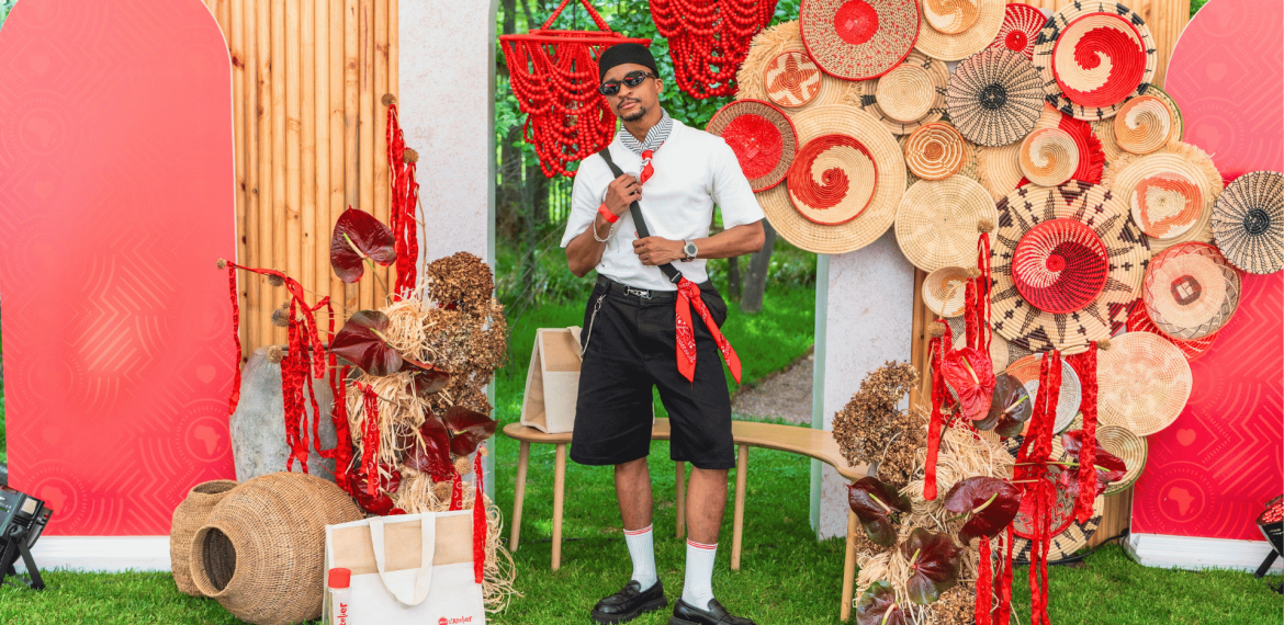 A man in sunglasses and casual clothes poses among woven baskets, red bead strands, and decorative patterns with red accents, outdoors on green grass, with red screens and wooden panels in the background.