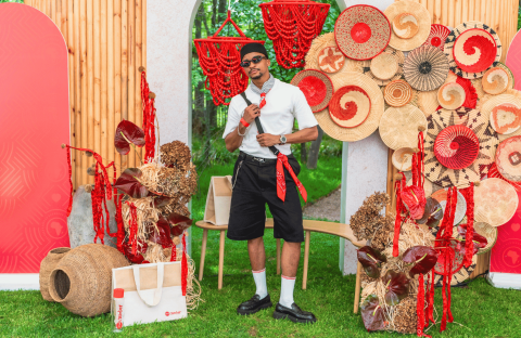 A man in sunglasses and casual clothes poses among woven baskets, red bead strands, and decorative patterns with red accents, outdoors on green grass, with red screens and wooden panels in the background.