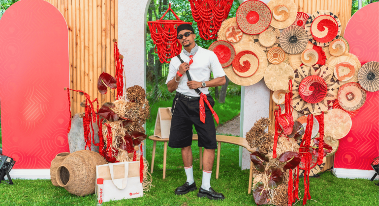 A man in sunglasses and casual clothes poses among woven baskets, red bead strands, and decorative patterns with red accents, outdoors on green grass, with red screens and wooden panels in the background.
