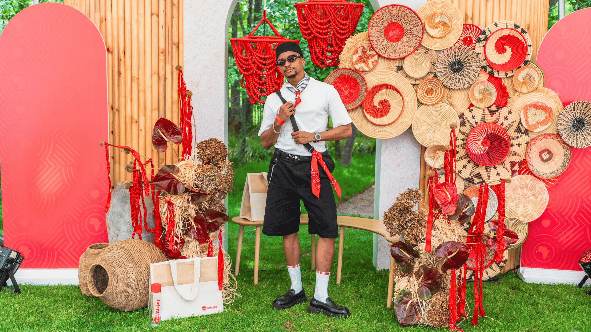 A man in sunglasses and casual clothes poses among woven baskets, red bead strands, and decorative patterns with red accents, outdoors on green grass, with red screens and wooden panels in the background.