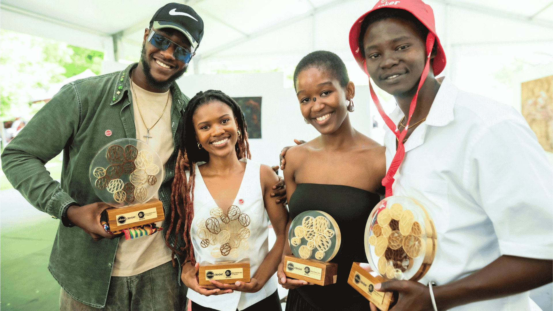 Four young adults stand side-by-side, smiling, each holding a round, abstract trophy with a wooden base at an Absa LAtelier art event. They are dressed in stylish, casual clothing including a cap and bucket hat.