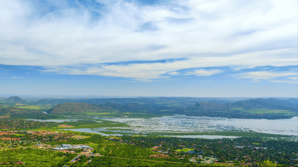 An aerial view of a sprawling landscape featuring a large lake with a grassy bank, surrounding greenery, residential areas, and distant mountains under a cloudy blue sky.