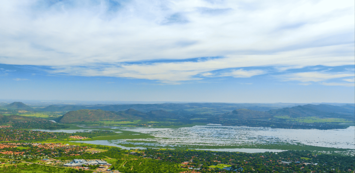 An aerial view of a sprawling landscape featuring a large lake with a grassy bank, surrounding greenery, residential areas, and distant mountains under a cloudy blue sky.