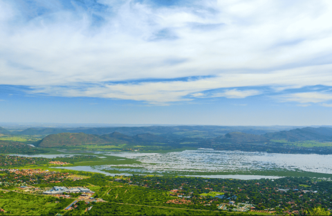 An aerial view of a sprawling landscape featuring a large lake with a grassy bank, surrounding greenery, residential areas, and distant mountains under a cloudy blue sky.