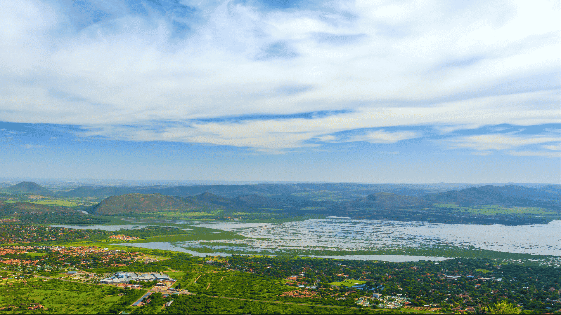 An aerial view of a sprawling landscape featuring a large lake with a grassy bank, surrounding greenery, residential areas, and distant mountains under a cloudy blue sky.