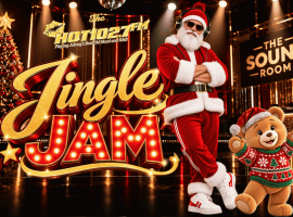 Santa Claus wearing headphones and a teddy bear in a festive sweater stand in a music studio in front of a Jingle Jam sign for Hot 102.7 FM.