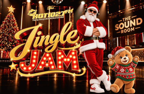 Santa Claus wearing headphones and a teddy bear in a festive sweater stand in a music studio in front of a Jingle Jam sign for Hot 102.7 FM.
