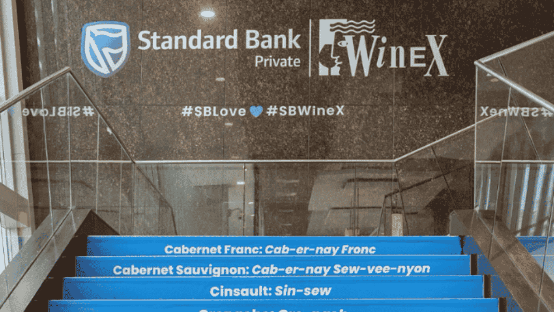 Staircase with blue treads listing wine names and pronunciations, beneath a wall featuring Standard Bank Private and WinEx logos with the hashtags #SBLove and #SBWineX.