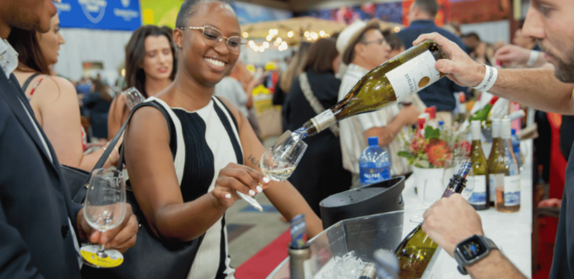 A server pours white wine into a glass held by a smiling woman at a crowded event.