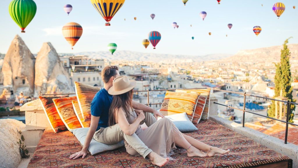 A couple sits on a patterned rug on a travel-inspired rooftop terrace overlooking a town in Cappadocia, with many hot air balloons floating in the sky above.