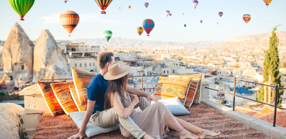 A couple sits on a patterned rug on a travel-inspired rooftop terrace overlooking a town in Cappadocia, with many hot air balloons floating in the sky above.