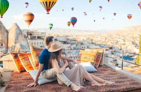 A couple sits on a patterned rug on a travel-inspired rooftop terrace overlooking a town in Cappadocia, with many hot air balloons floating in the sky above.