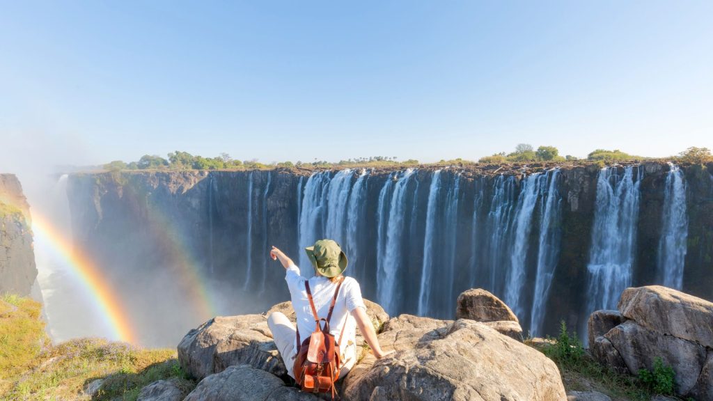A person wearing a sun hat and backpack sits on a rocky cliff, pointing toward a large waterfall with a rainbow in the mist during their travels.
