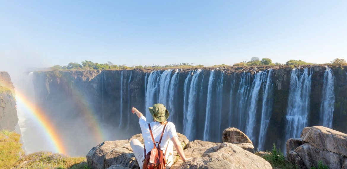 A person wearing a sun hat and backpack sits on a rocky cliff, pointing toward a large waterfall with a rainbow in the mist during their travels.