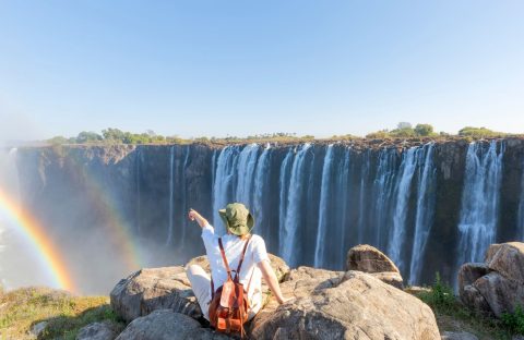 A person wearing a sun hat and backpack sits on a rocky cliff, pointing toward a large waterfall with a rainbow in the mist during their travels.