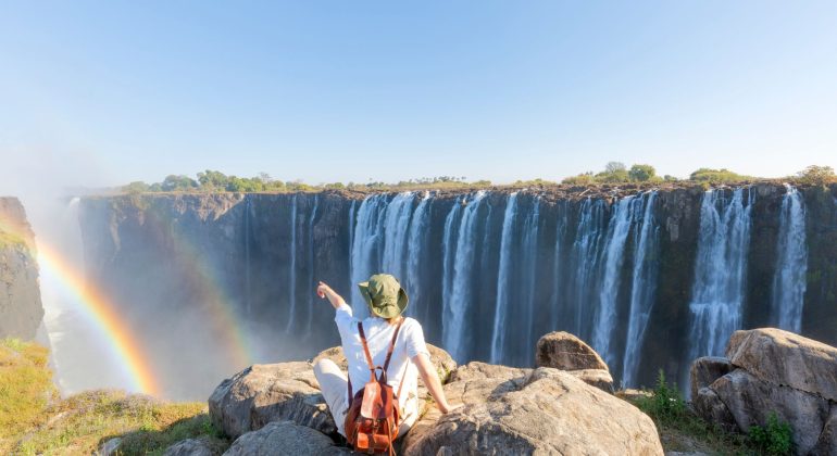 A person wearing a sun hat and backpack sits on a rocky cliff, pointing toward a large waterfall with a rainbow in the mist during their travels.