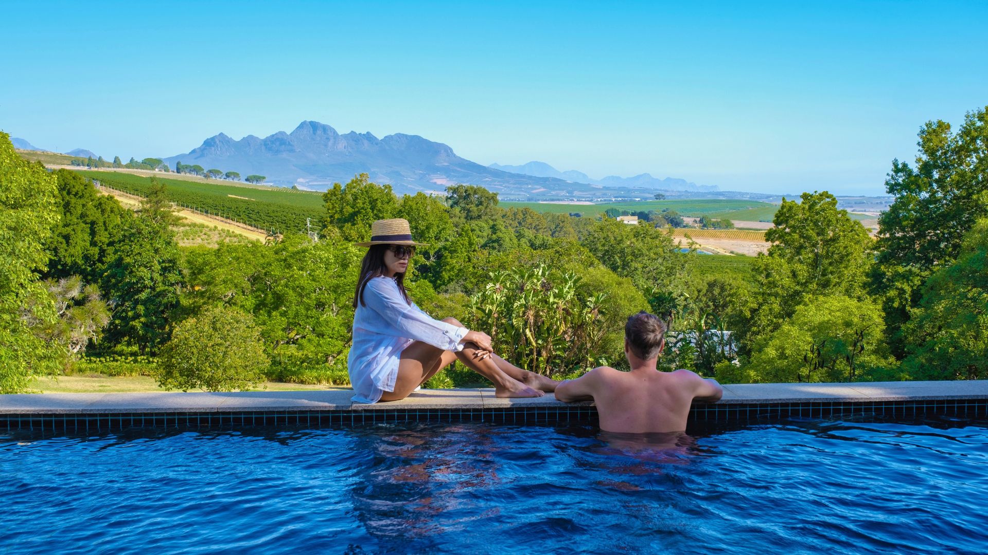 A woman in a sun hat and a man sit on the edge of an infinity pool overlooking a travel destination featuring a valley with vineyards and mountains.