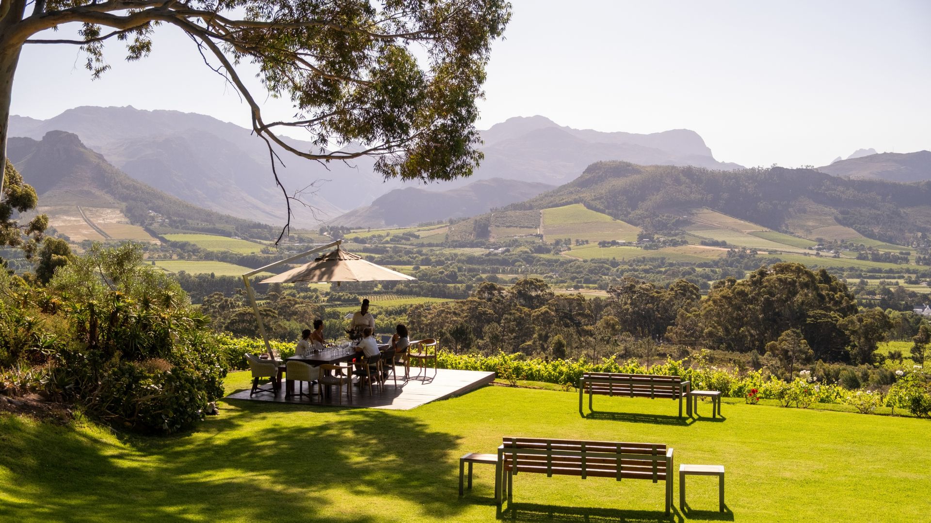 People enjoy a travel dining experience at an outdoor table on a wooden deck overlooking a vineyard and mountain range.
