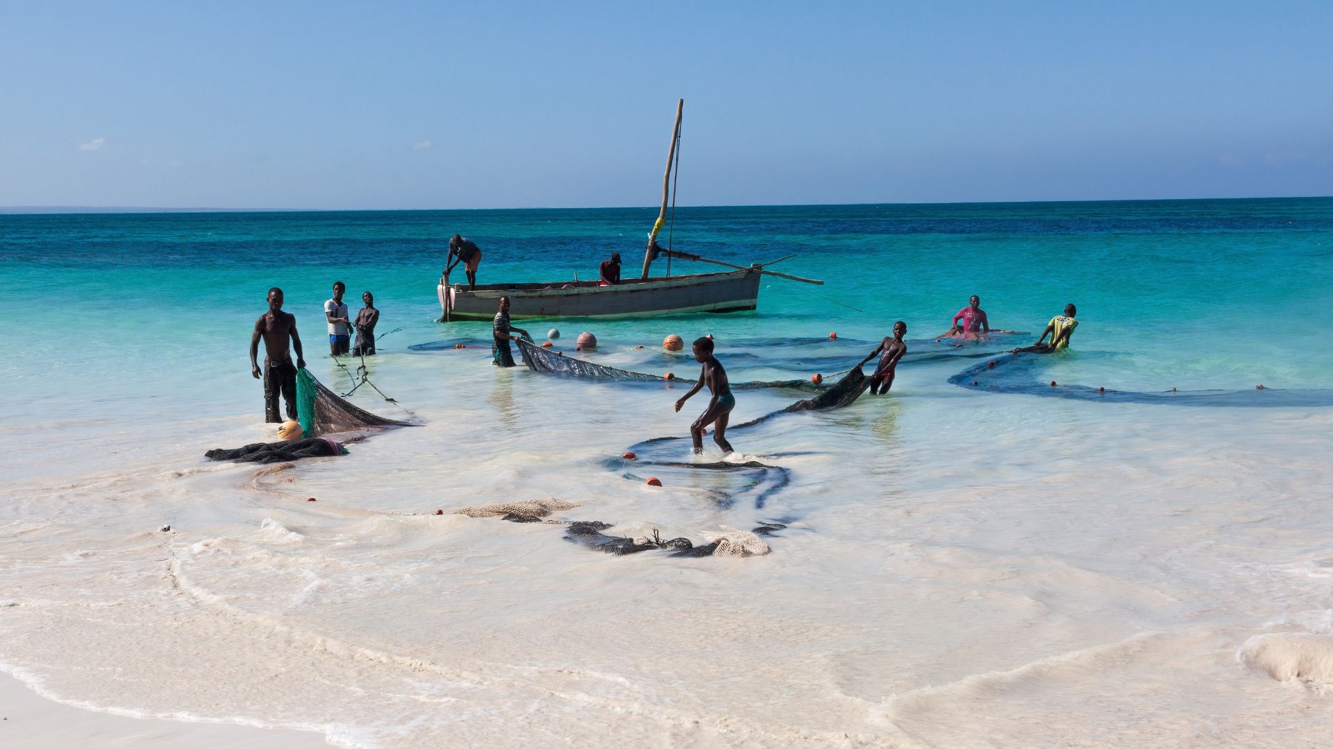 A group of people on a travel excursion stand in shallow turquoise water pulling a long fishing net, with a wooden boat moored nearby.