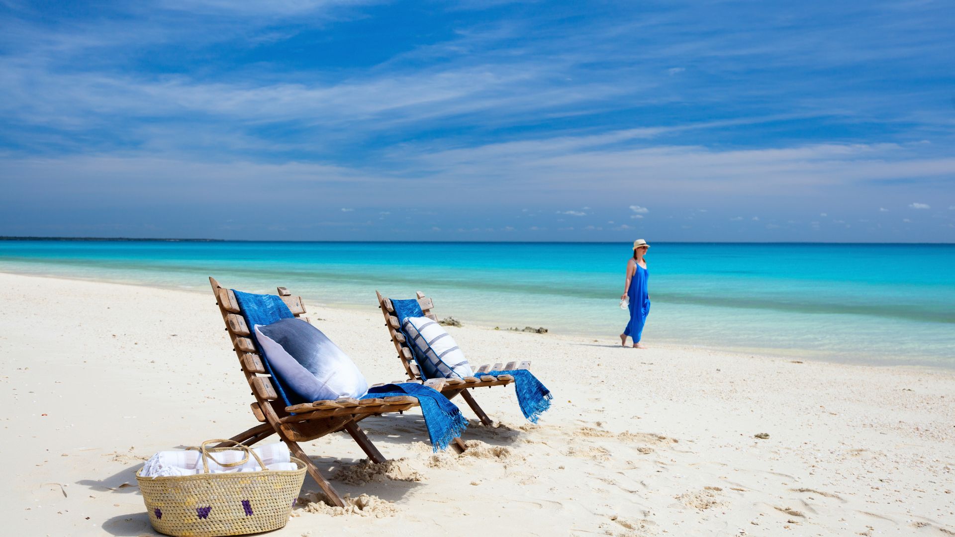 Two wooden beach chairs with pillows and blue blankets sit on white sand as a woman in a blue dress walks along the shoreline of a turquoise sea, a serene travel scene.