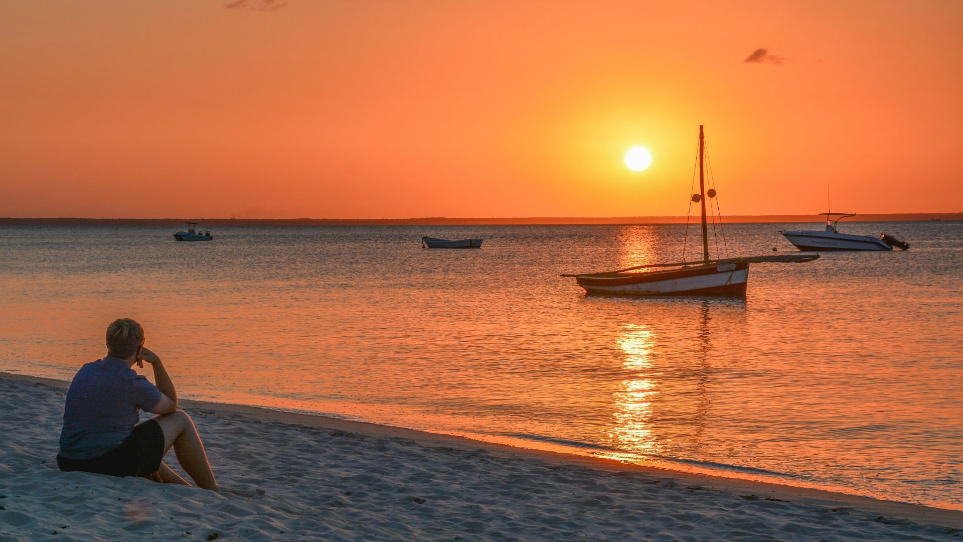 A person sits on a sandy beach during a bright orange travel sunset, looking out at boats on the water.