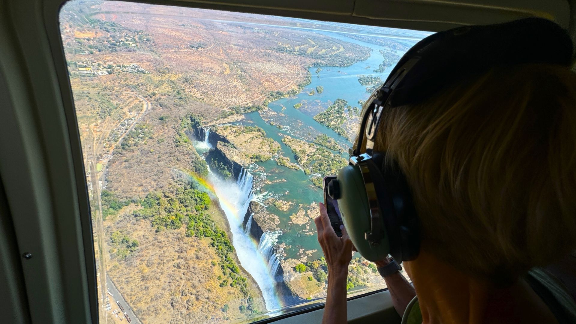 A passenger wearing noise-canceling headphones captures a travel photo of Victoria Falls and a rainbow from a helicopter.