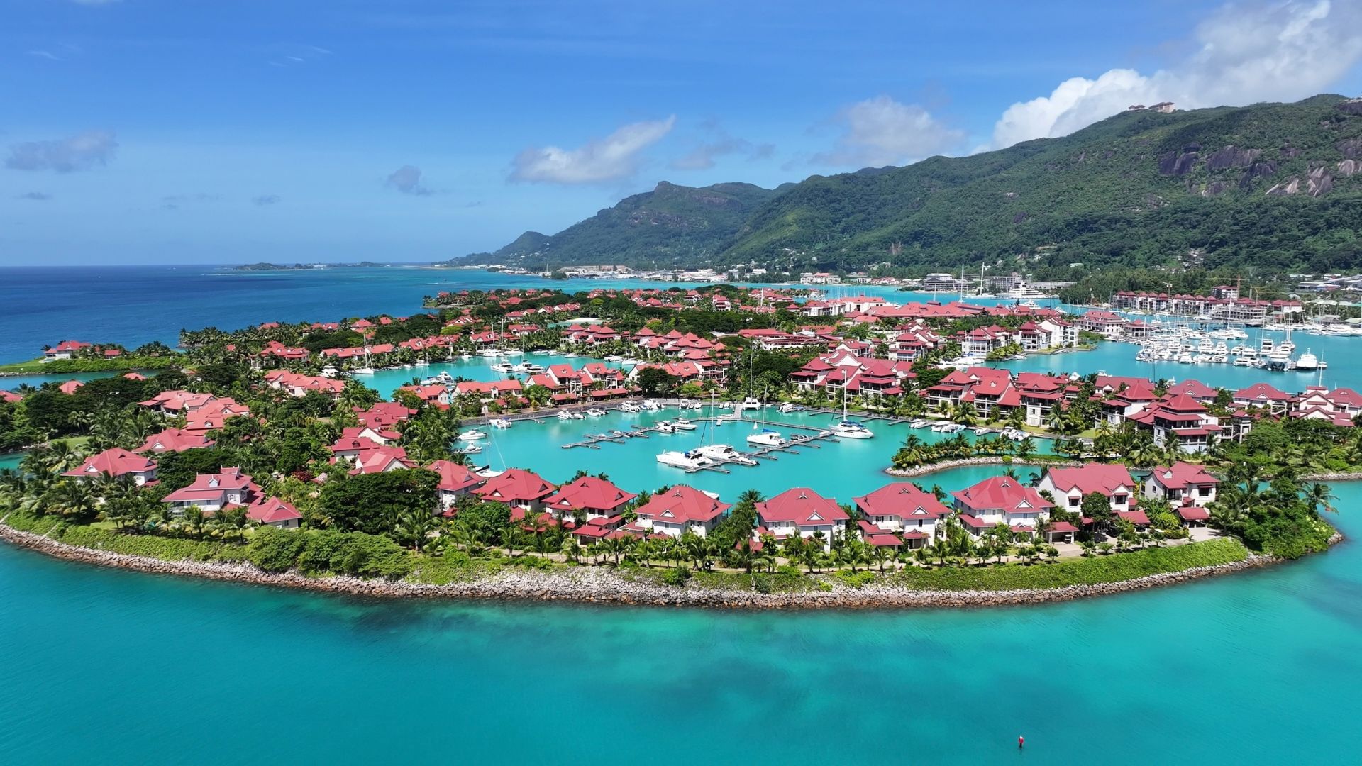 Aerial view of a luxury residential marina with red-roofed villas surrounding a turquoise travel destination bay filled with boats, backed by lush green mountains.