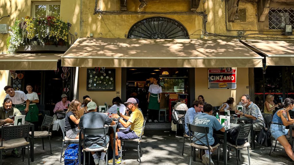 Travelers dine at outdoor tables in front of a cafe with a Cinzano sign and a waiter standing in the doorway.