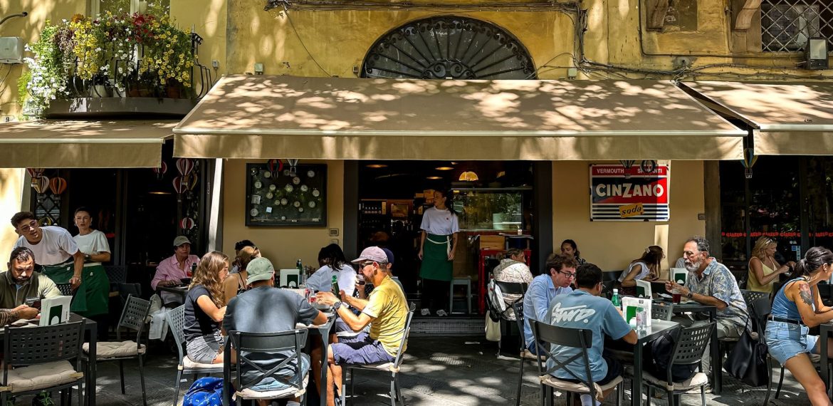 Travelers dine at outdoor tables in front of a cafe with a Cinzano sign and a waiter standing in the doorway.
