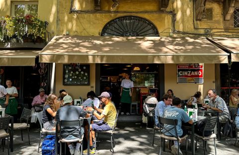 Travelers dine at outdoor tables in front of a cafe with a Cinzano sign and a waiter standing in the doorway.